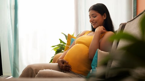 Young South Asian woman sitting on a couch looking happily at her pregnant belly