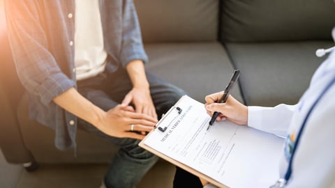A doctor writing on a clipboard in front of a patient.