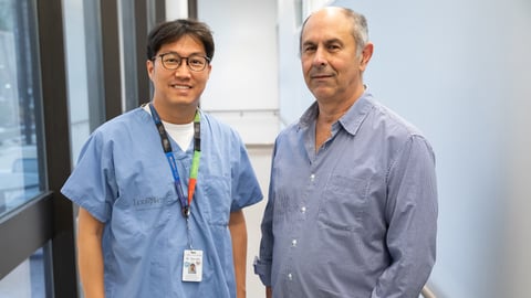 A man in physician's blue scrubs and a man in a patterned button down shirt stand in a hallway.