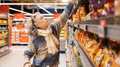 An older white women in the grocery store pulling a bag of chips from a high shelf