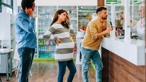 A lineup of three people at pharmacy window, each looking impatient, including a pregnant woman.