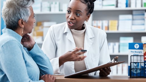 A pharmacist holding a clipboard speaks with a patient over the pharmacy counter.