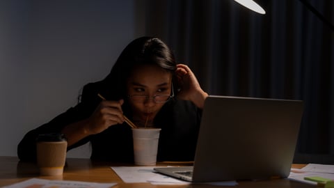 Young woman working late eating noodles while looking at her laptop