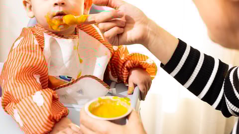 Baby with an orange outfit eating baby food and spitting a lot of it out