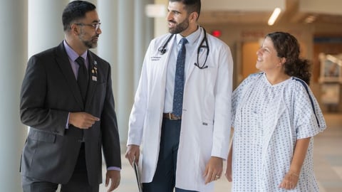 Two male physicians, one in a suit and one in a lab coat, and a young woman patient in a hospital gown, walk while talking in a hallway