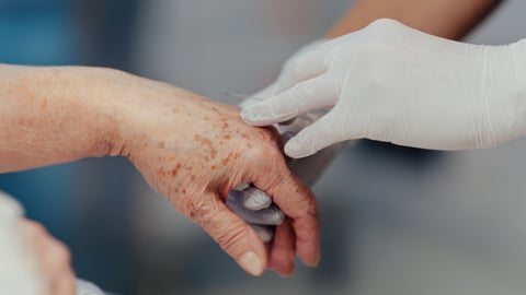 Glove hand of a nurse holding the hand of an older patient receiving MAiD