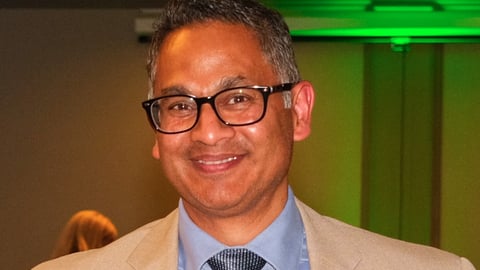 Glenn Rodrigues, in a brown suit smiling for a photo