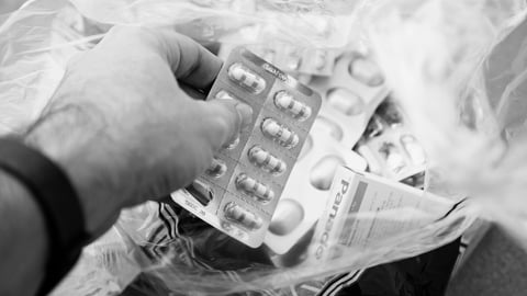 Black and white photo of a white man's hand reaching into a plastic bag filled with prescription drug tablets