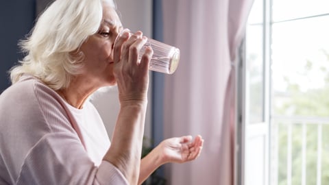 Older white woman in a pink shirt taking a pill with water