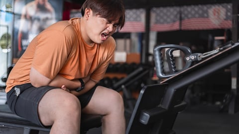 Young Asian man in orange shirt at gym, grimacing after over-exercising, in pain