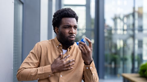 Young Black man with a yellow shirt clutching his chest and holding an asthma inhaler