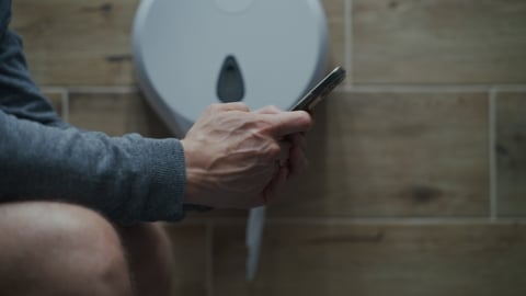 A man uses his phone while sitting on the toilet.