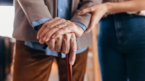 An older man's hands on a cane, being helped along by a younger person