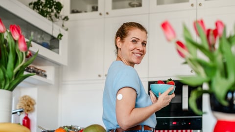 Happy young white woman in her kitchen with a bowl of fruit and a small wearable health monitor on her arm
