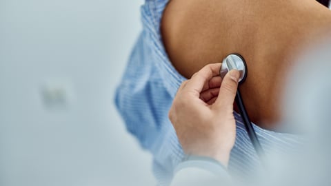 A physician places a stethoscope on a patient's exposed back.