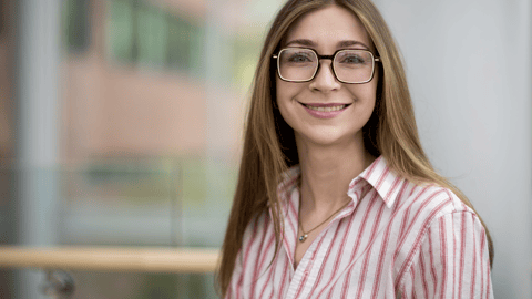 A young white woman with long brown hair wearing glasses and a pink shirt looks at the camera.
