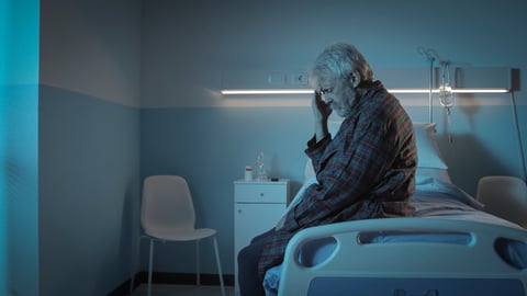 A senior with his hand on his head while sitting on a hospital bed.