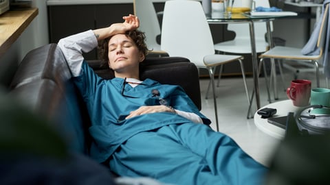 A woman doctor in blue scrubs taking a nap on a break room couch