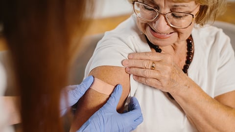 A healthcare provider putting a bandage on the upper arm of a smiling older woman with glasses after a vaccination.