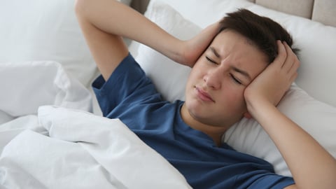 A young teenage boy holding his head in bed with a migraine