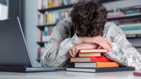 A student at his desk with a laptop, sleeping with his head on top of a stack of books.