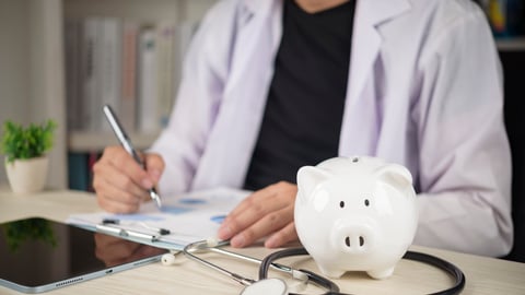 A doctor fills out paperwork next to a piggybank.