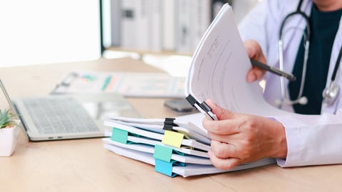 A doctor examines stacks of paper representing medical records and paperwork.