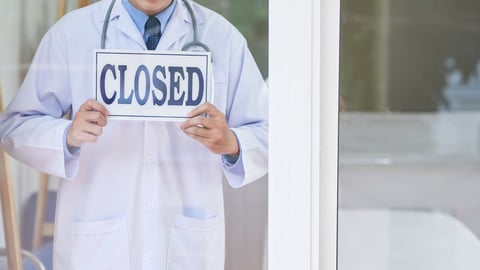 Anonymous guy in medical uniform holding sign with closed writing while standing behind glass in office; Shutterstock ID 1171458436