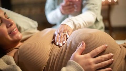 Cropped image of smiling pregnant woman and midwife at home. Woman in casual clothes lying on bed, midwife holding hand. Pregnancy, medicine, home birth concept; Shutterstock ID 2145598557
