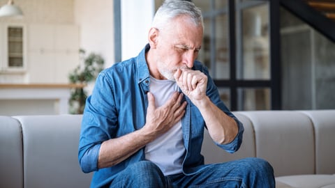 Senior man sitting on sofa at home and holding hand on chest. Male having asthma attack, difficulties with breathe, feeling severe pain or dyspnea; Shutterstock ID 2420095589