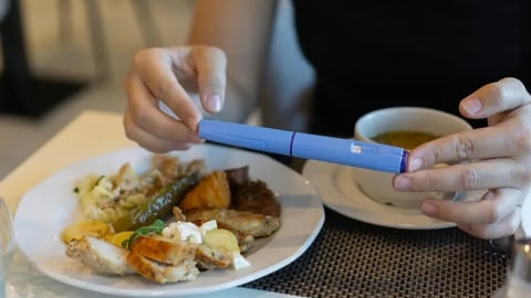 Close-up of hands holding an Ozempic injection pen while sitting at a table with a plate of food and a cup of soup; Shutterstock ID 2675228061