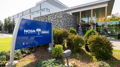 A blue sign saying NOSM is outside a modern education building about four stories high.