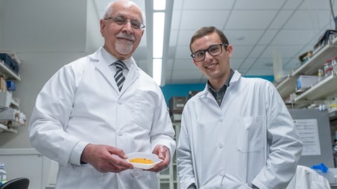 Two men in labcoats stand in a lab holding a bowl of yellow-orange berries.