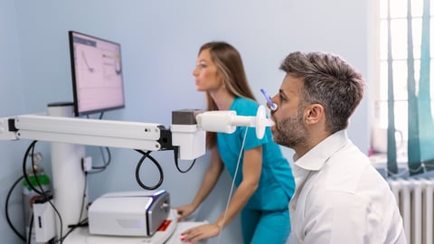 man undergoing spirometry with tube in mouth and plug on nose, famale technician in background