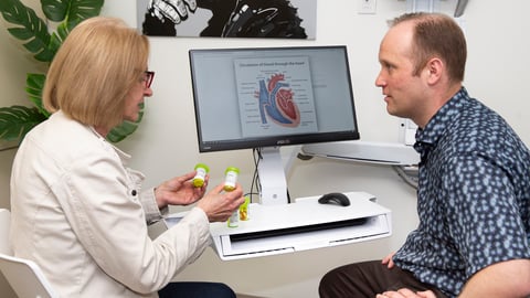 A pharmacist and a patient talking with a computer showing an image of a heart in the background