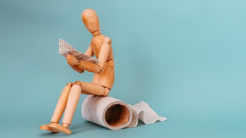 Wooden figure sitting on roll of toilet paper, reading newspaper