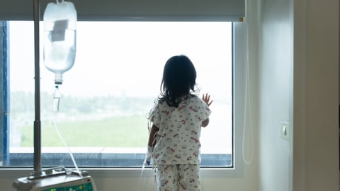 Child wearing IV tube is standing and looking out the window in hospital