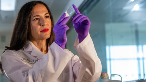 A woman with long brown hair and bold lipstick looks at a microscope slide while wearing a lab coat and purple gloves.