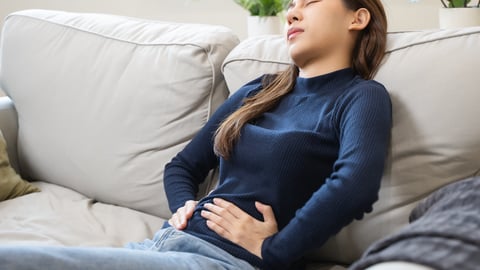 Young Asian woman on beige couch grimacing and holding lower abdomen