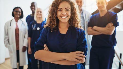 Young female doctor smiling while standing in a hospital corridor with a diverse group of staff in the background; Shutterstock ID 1208448832