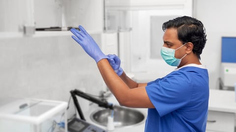 healthcare, profession and medicine concept - indian doctor or male nurse in blue uniform and face protective medical mask putting surgical gloves on over laboratory or hospital background; Shutterstock ID 1928391047