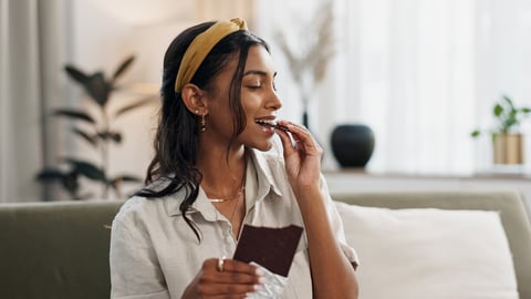 Home, relax and woman with chocolate bar, smile and eating with candy, hungry and dessert. Person, apartment or girl on a sofa, candy or sweet treats with a snack, nutrition or diet plan with cacao; Shutterstock ID 2377580049