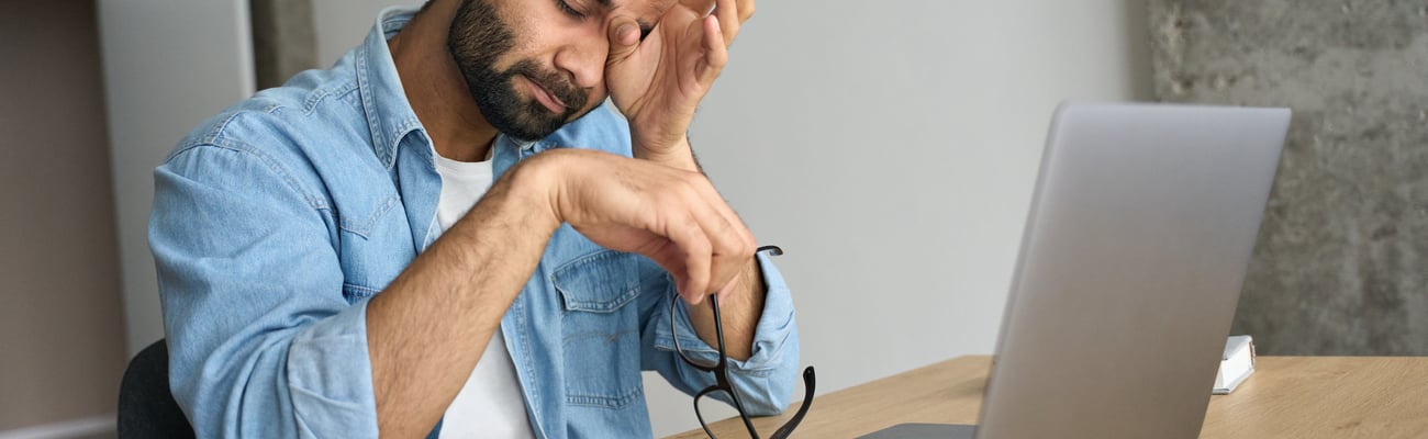 A man with a beard sits in front to a laptop computer wiping his eyes