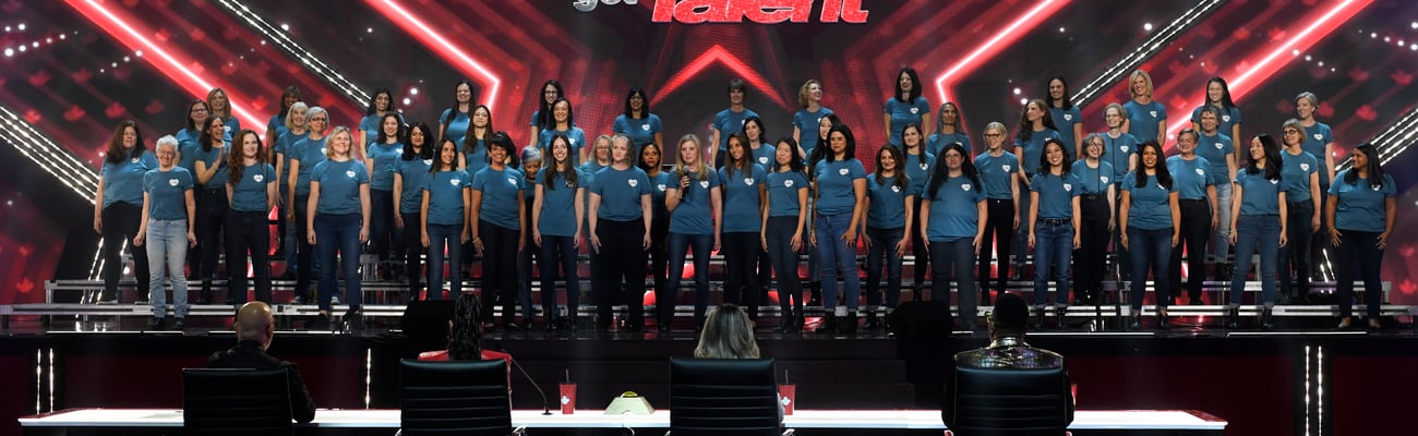 Voices Rock Medicine choir on stage at Canada's Got Talent with more than 50 members wearing jeans and blue t-shirts standing on a stage with red lights spelling out Canada's Got Talent