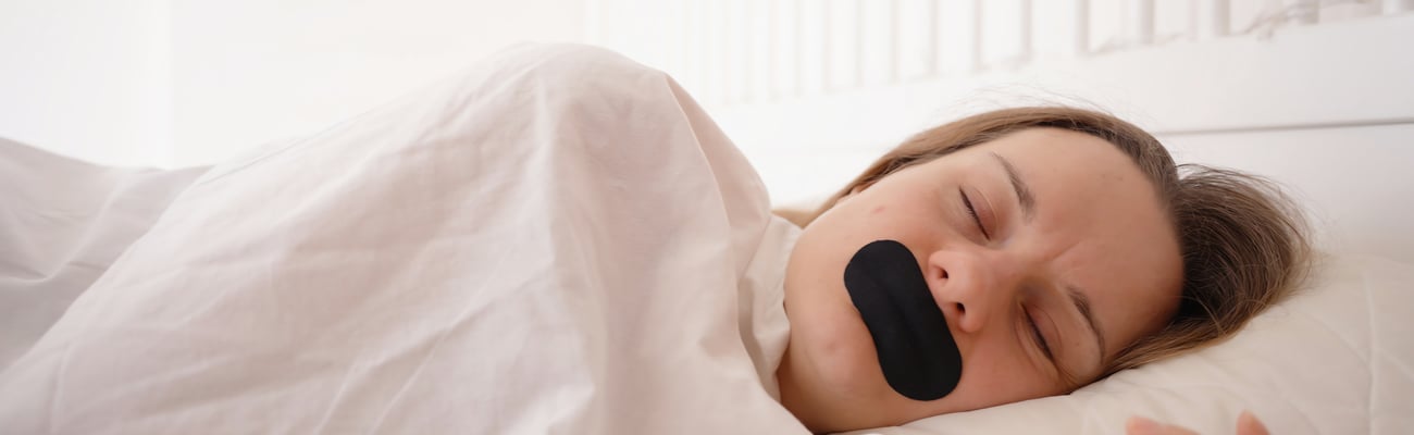 Woman sleeping on side under white blanket with black mouth tape in bright bedroom