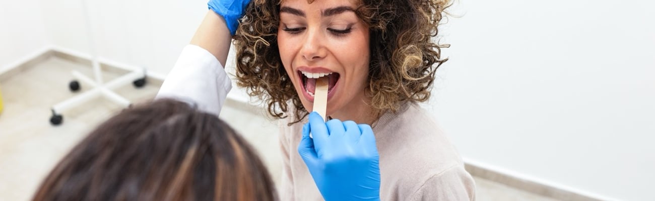 Overhead view of doctor examining woman patient's throat using a wooden tongue depressor and wearing blue medical gloves.