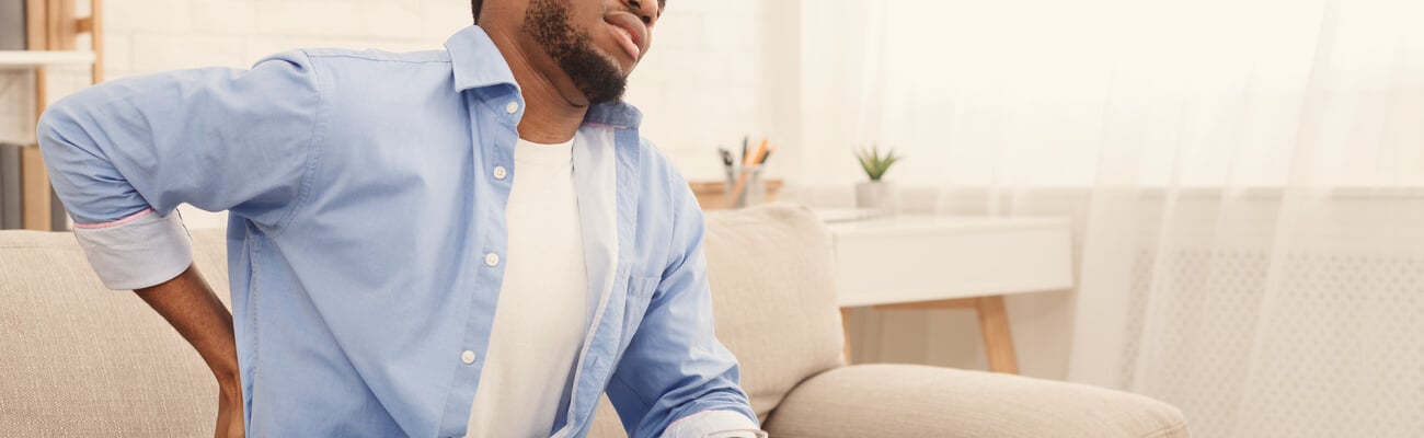 Young african-american man with back pain, pressing on hip with painful expression, sitting on sofa at home with glass of water, copy space; Shutterstock ID 1286261095