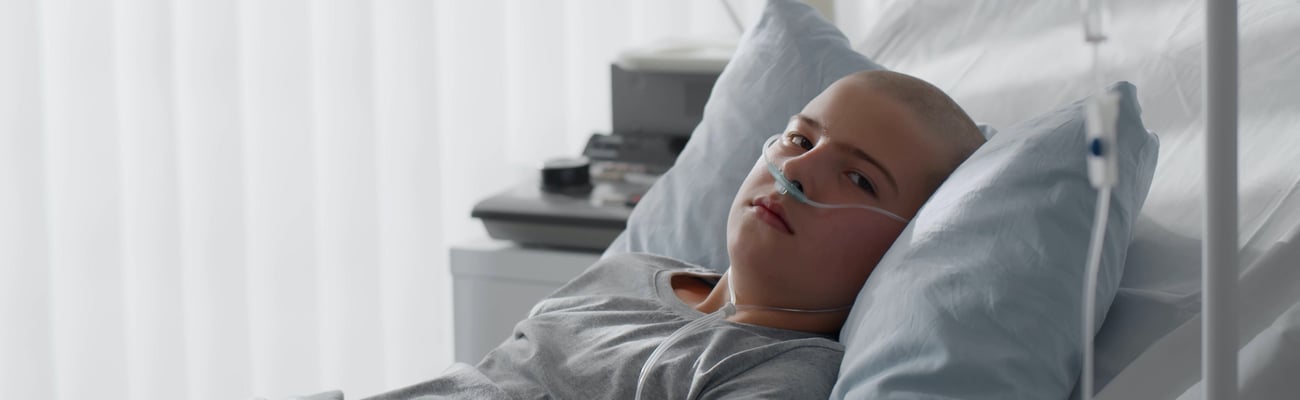 boy/teen in hospital bed, no hair, looking at camera, serious expression