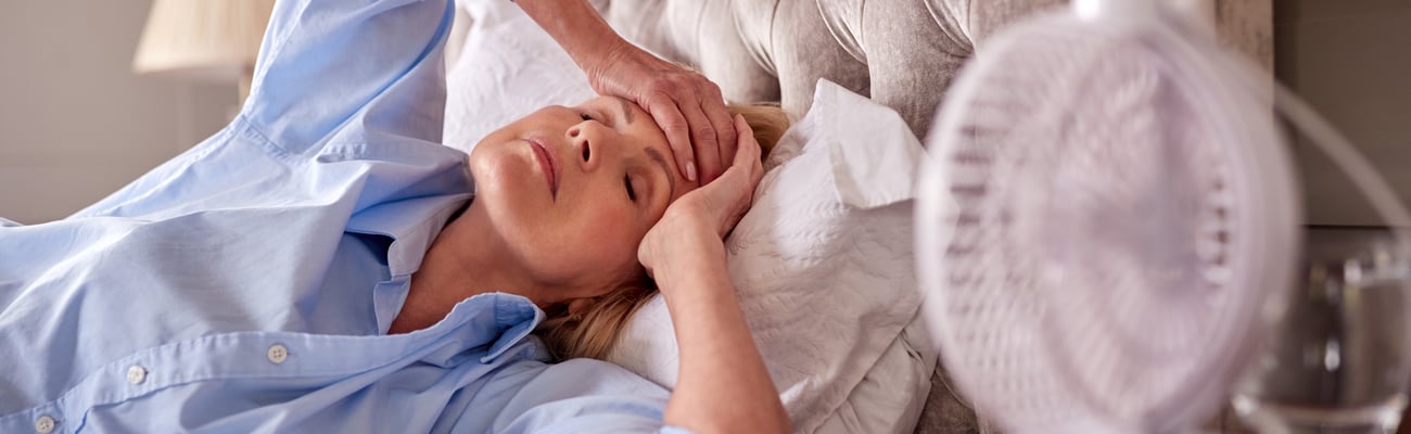 Woman in bed holding forehead with eyes closed and fan on her, suggesting hot flush