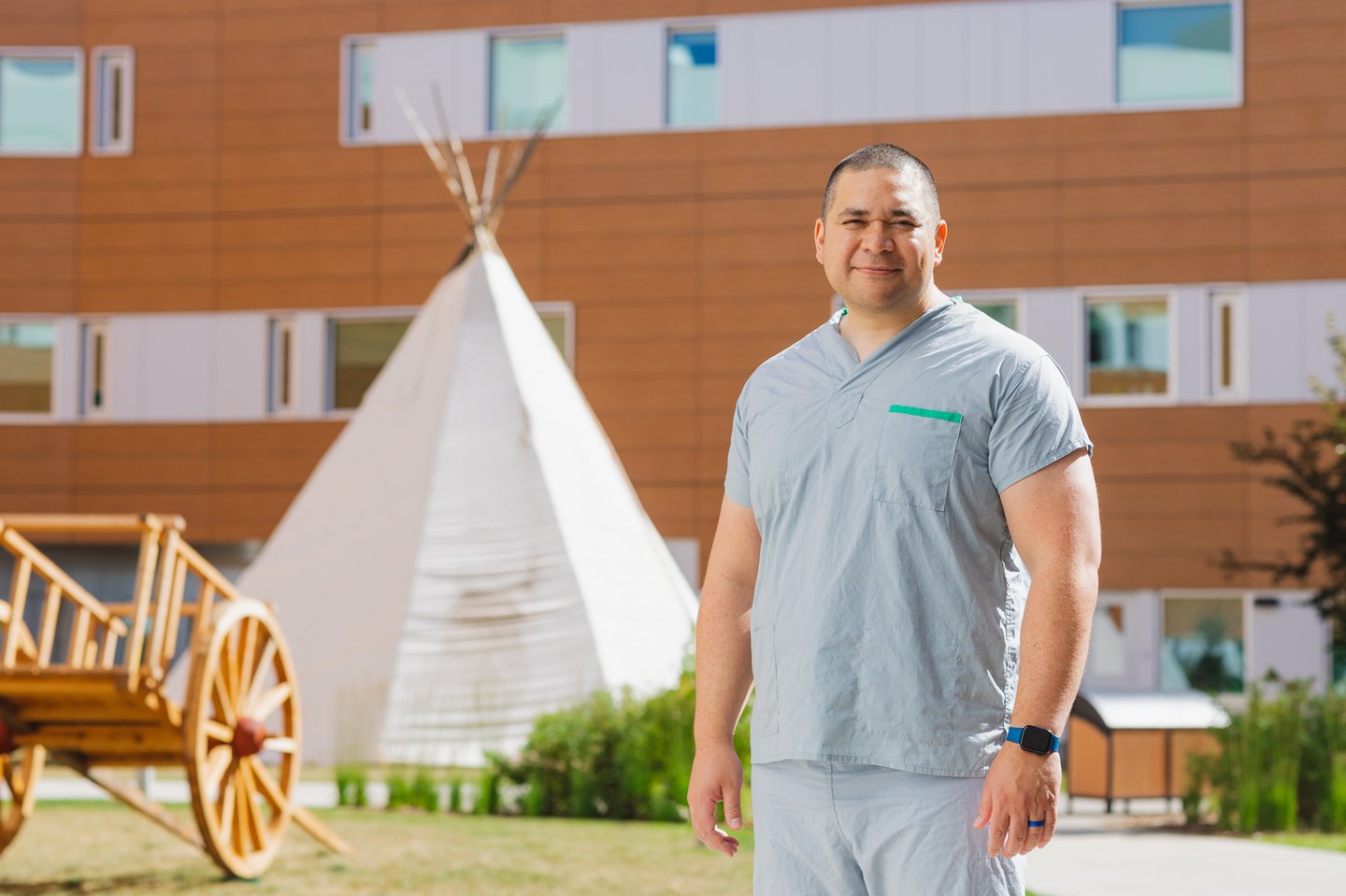 Tall man wearing scrubs outside a hospital in front of a traditional Indigenous structure